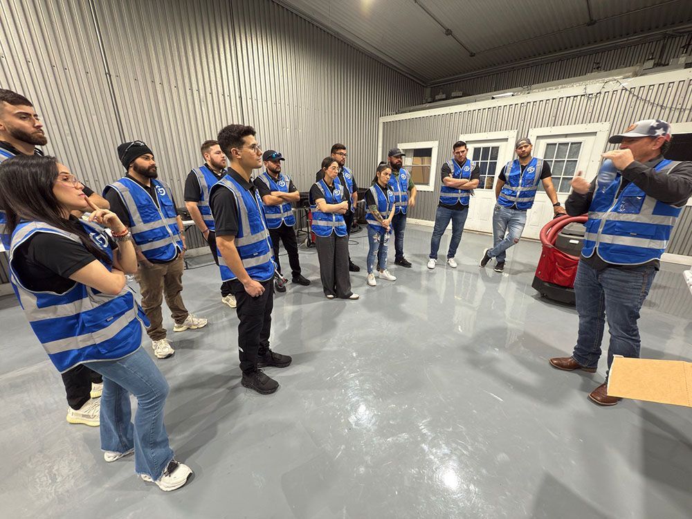 People in blue safety vests listen during a group briefing in a garage or warehouse.