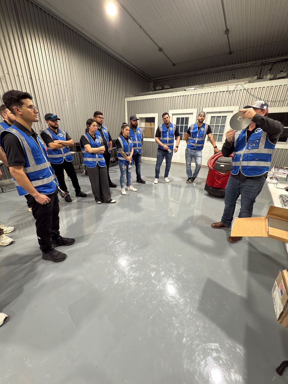People in blue safety vests stand in a warehouse, gathered in a circle during a briefing.