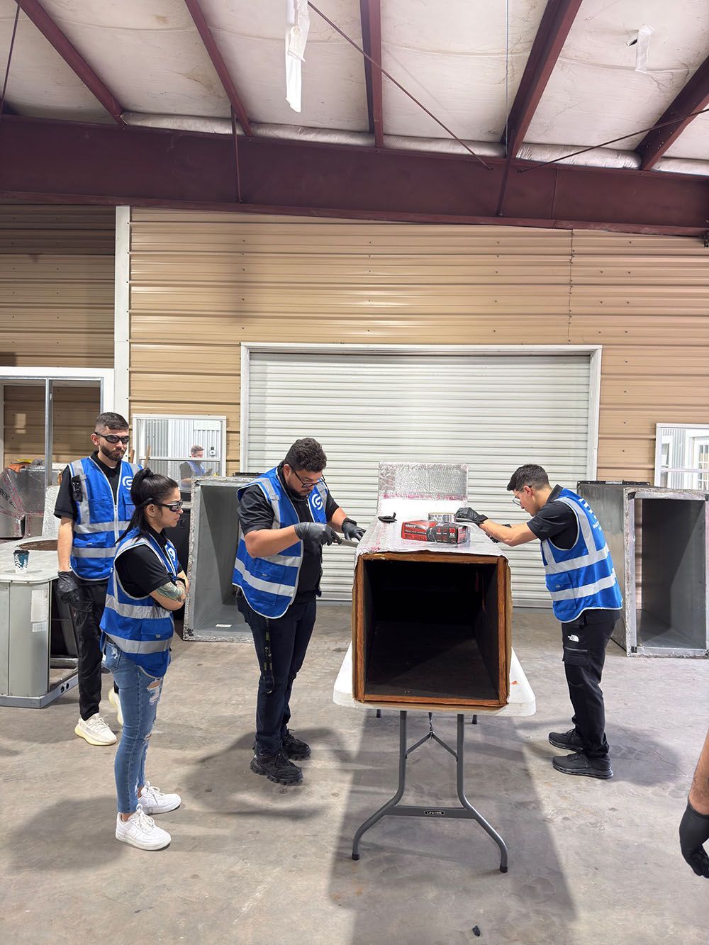 Four people in blue safety vests gather around a large wooden box in a warehouse.