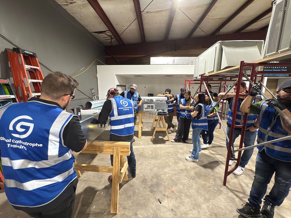 People in blue safety vests gather in a warehouse around wooden workstations and metal shelving.