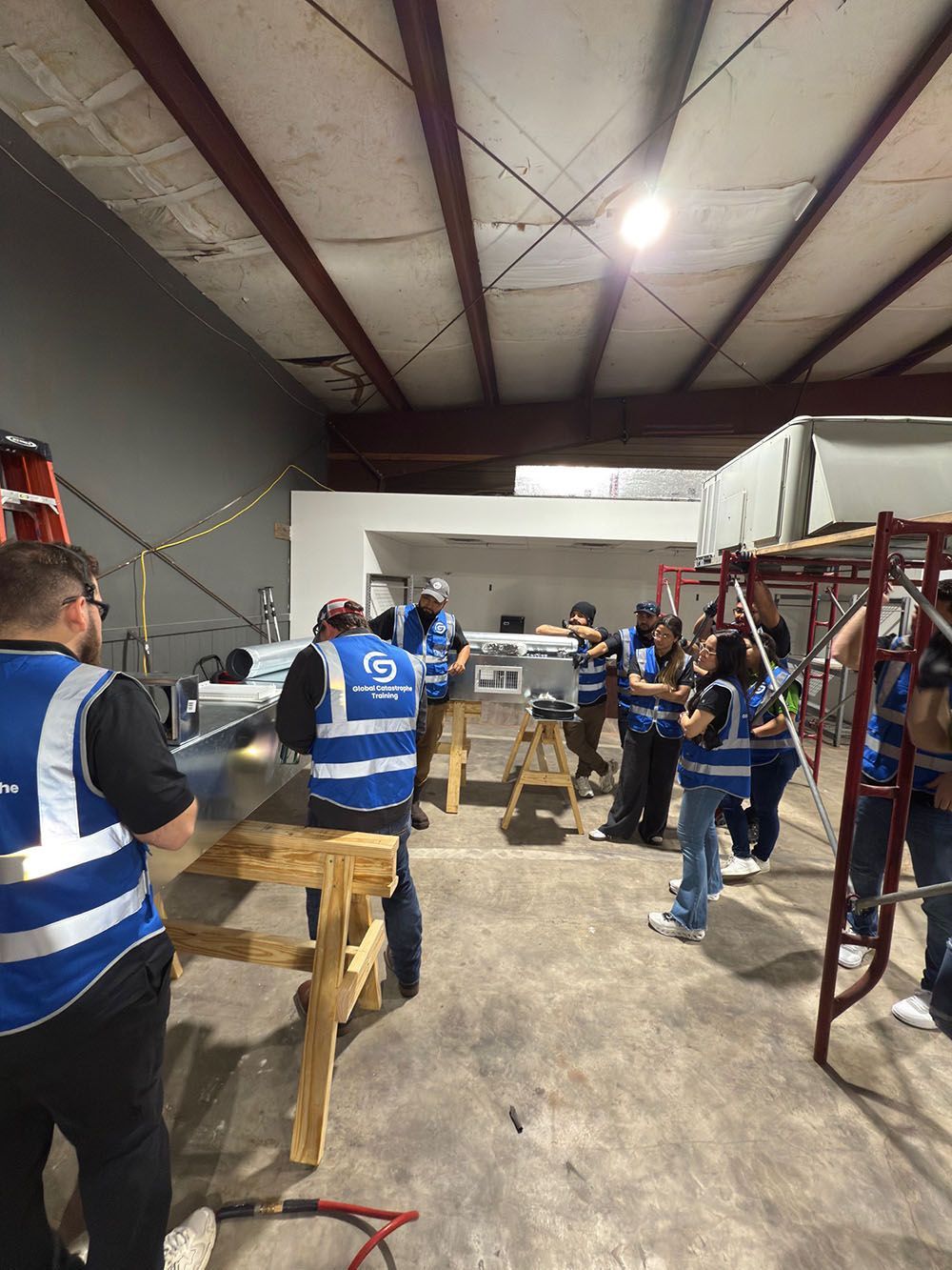 People in blue safety vests work around wooden benches in a warehouse workshop.