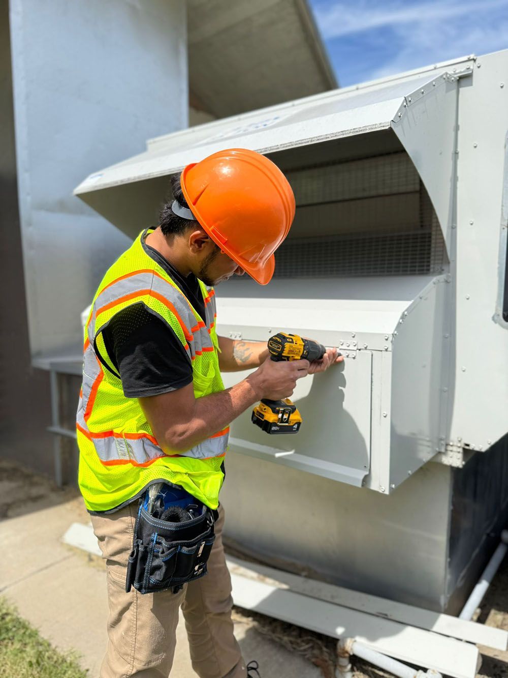 Worker in orange hard hat installing a metal vent on a rooftop with a power drill