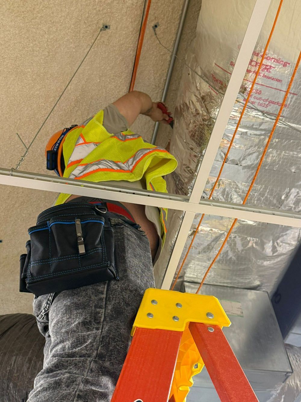 Worker in a yellow safety vest on a ladder, reaching toward ceiling insulation in a construction site.