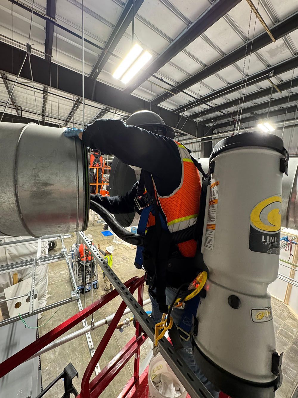Worker in orange safety vest inspecting large metal ductwork inside an industrial facility.