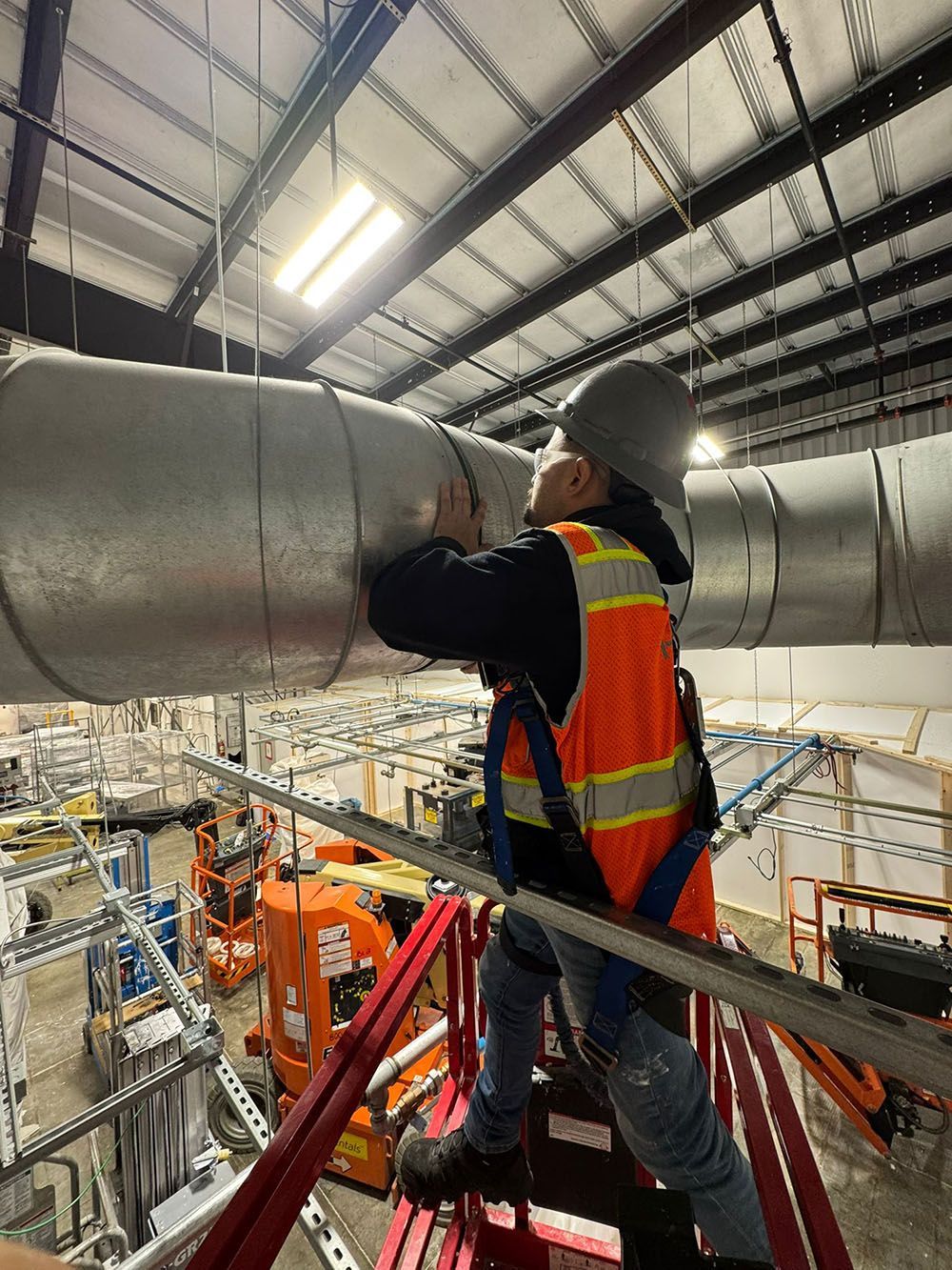 Worker in orange safety vest inspecting large silver ductwork on a red platform inside an industrial warehouse.