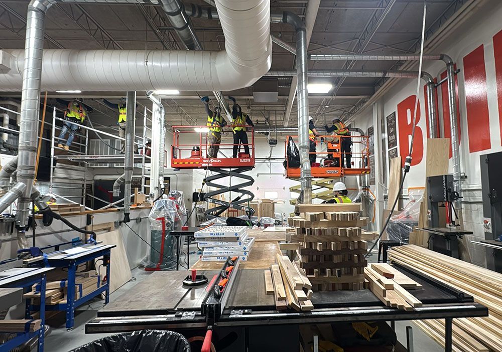 Industrial workshop with workers on lifts above a cluttered workbench and stacked lumber under bright lights