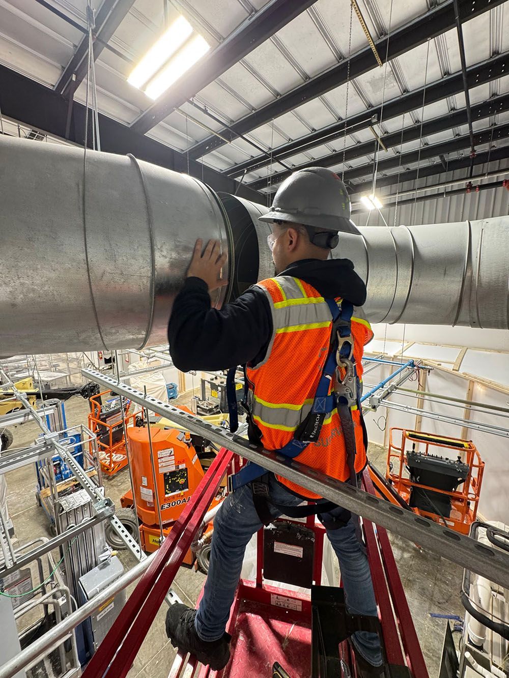Worker in hard hat and safety vest standing on a maintenance platform inside an industrial facility, inspecting ductwork.