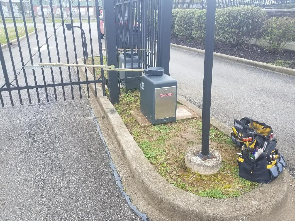 Black gate entrance beside a curb, with a small utility box and a tool bag on the grass.