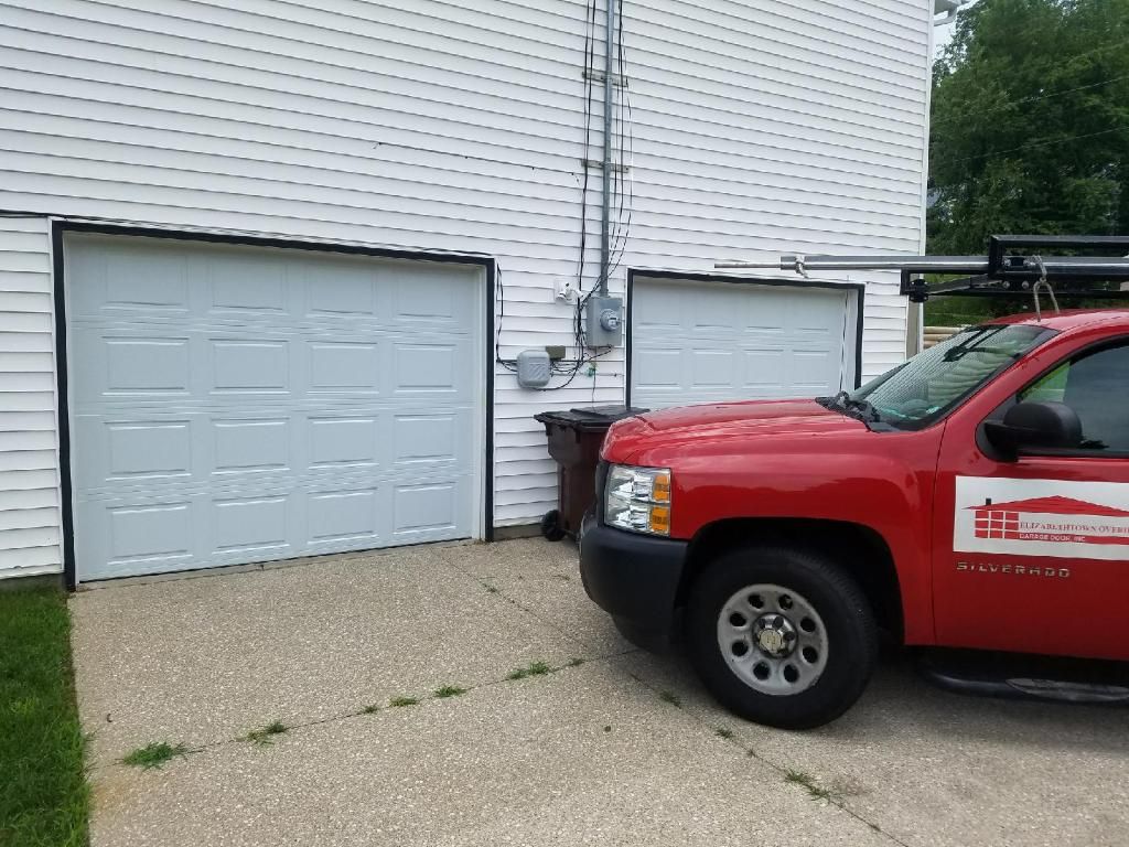 Red pickup truck parked beside two white garage doors on a gravel driveway.