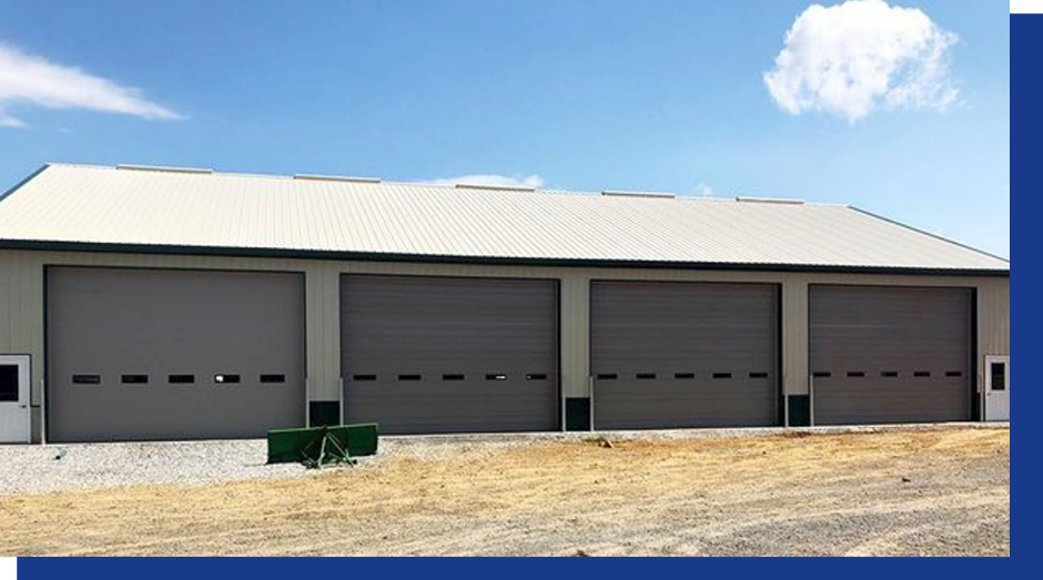 Large gray industrial building with four garage doors under a white roof on a dry lot