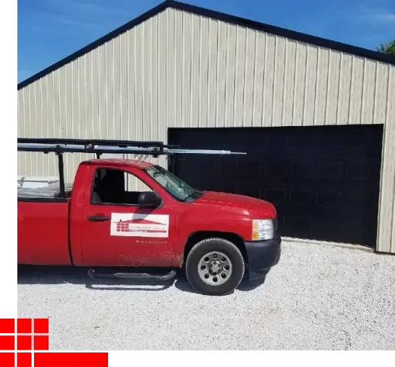 Red pickup truck parked beside a large beige metal garage on a gravel lot.