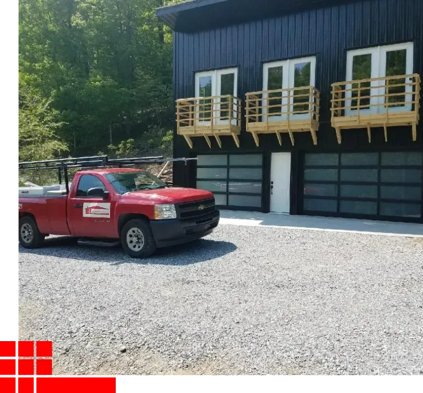 Red pickup truck parked beside a dark building with wooden balconies on a gravel lot, trees in the background