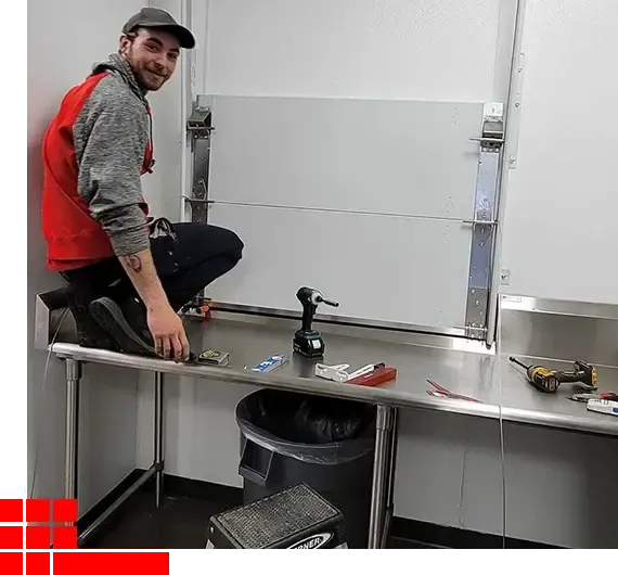 Man kneels on a metal workbench in a workshop, smiling beside tools and a trash bin.