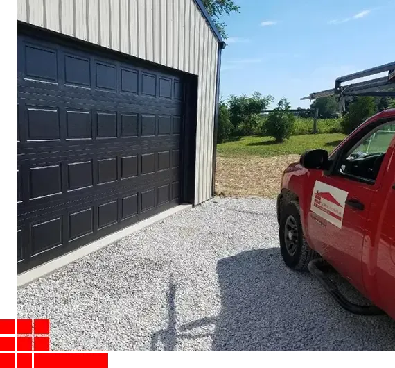 Red pickup truck beside a large black garage door on a gravel driveway near open fields.