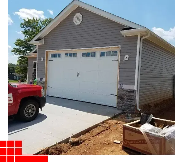 Front view of a gray garage with a white double door, driveway, and red truck under construction.