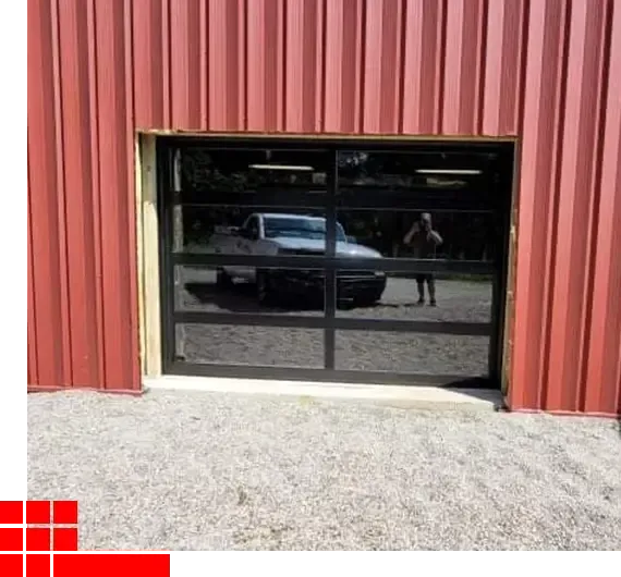 Red metal building with a large reflective black garage door and gravel driveway in front.