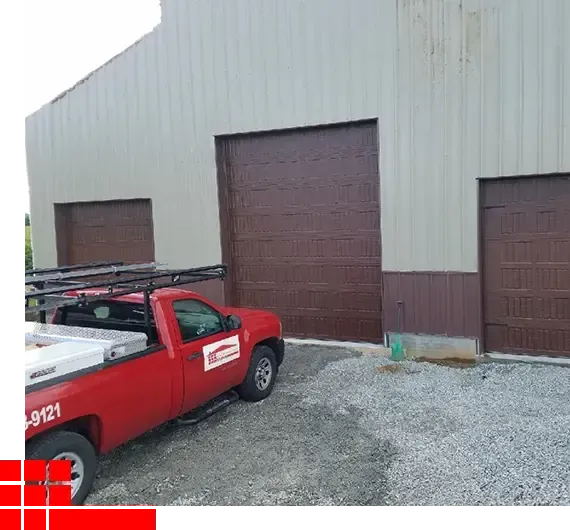 Red utility truck parked outside a beige metal warehouse with brown roll-up doors.