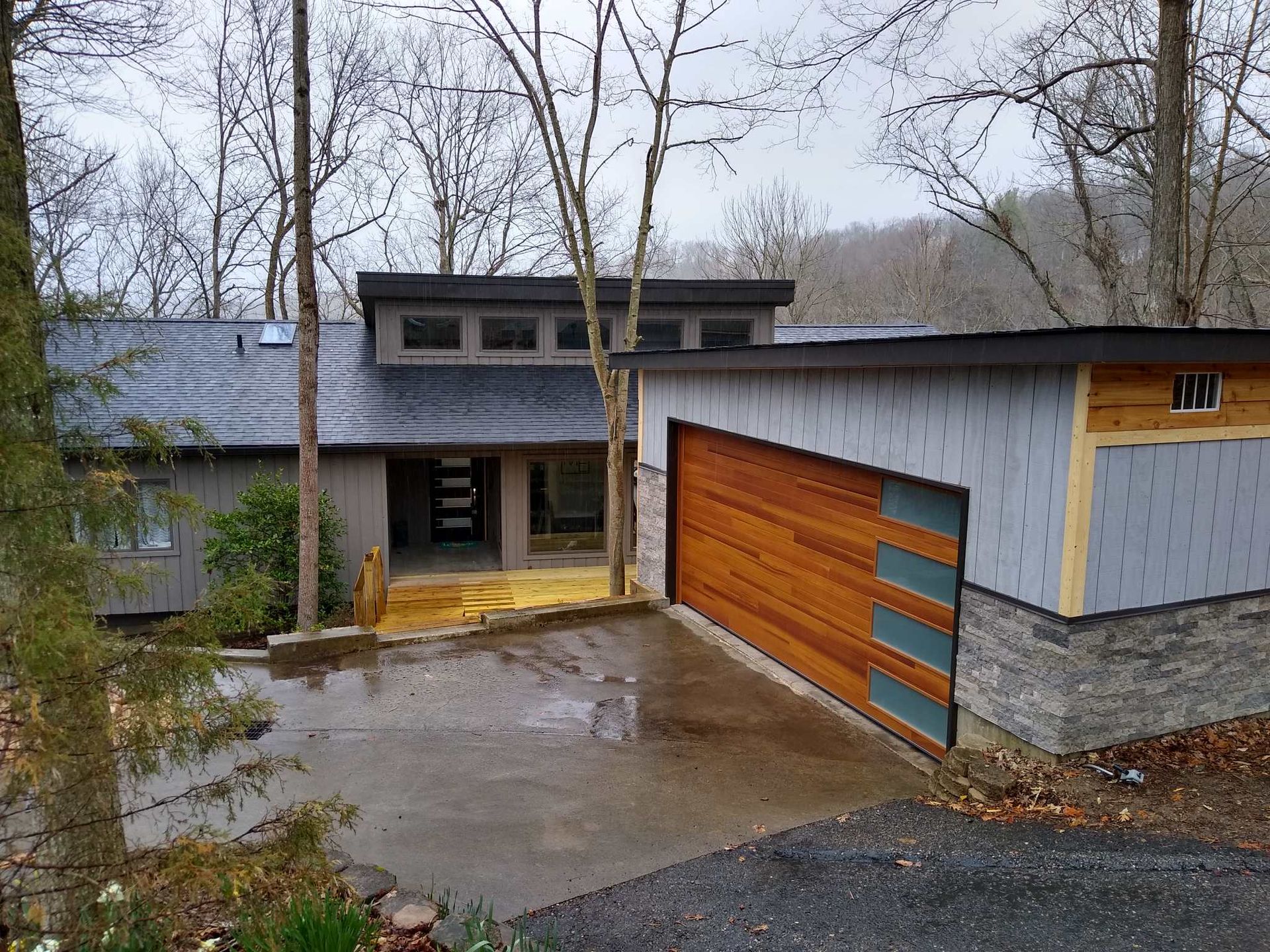 Modern house exterior with wet driveway, wood garage door, and leafless trees in front.