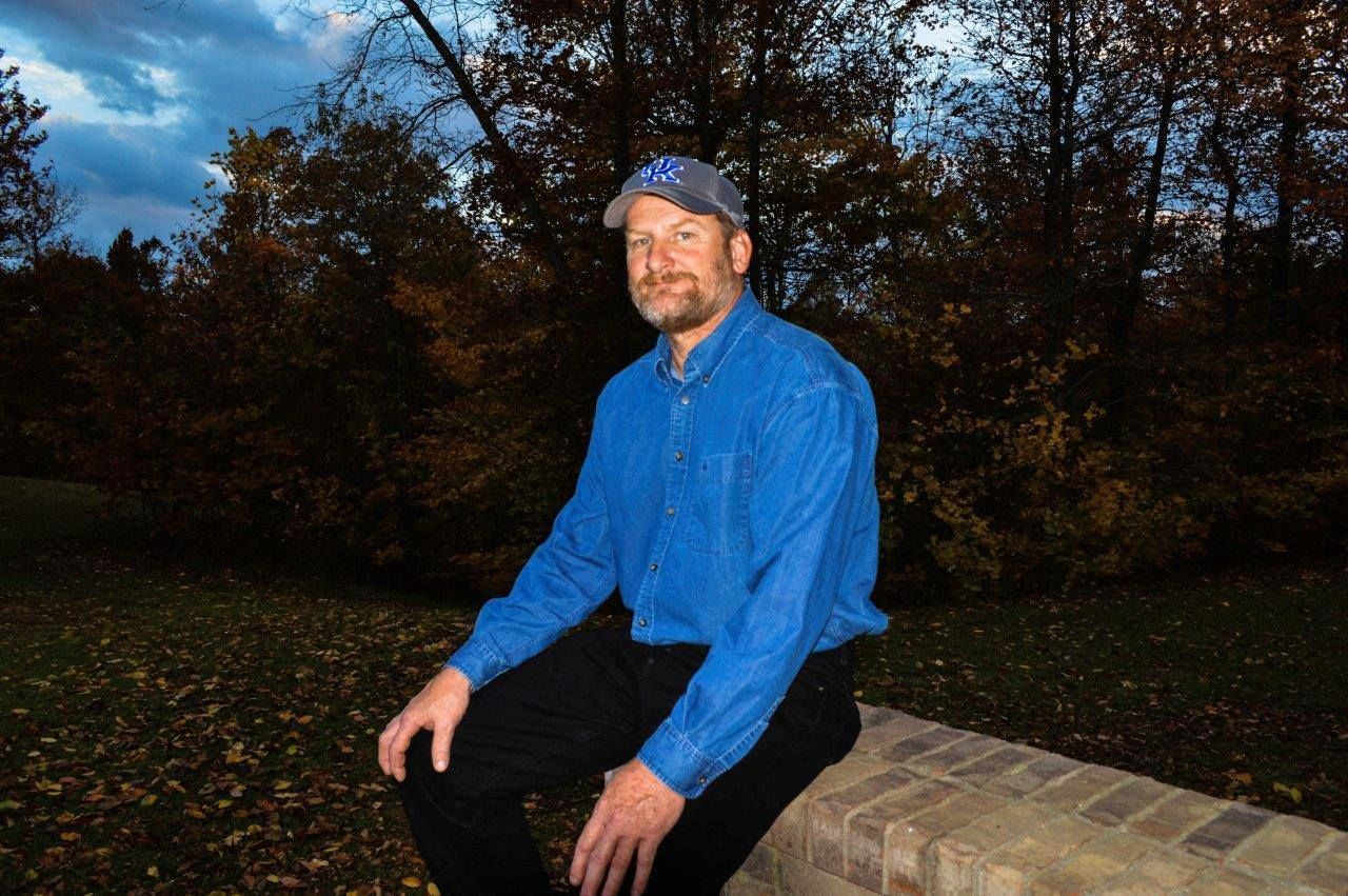 Man in a blue shirt sitting on a stone wall outdoors at dusk with trees in the background