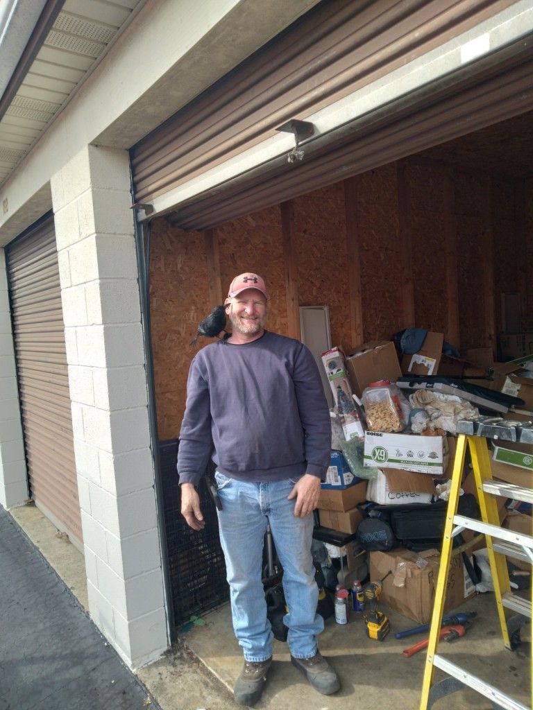 Man standing in an open garage beside a ladder and piles of stored items