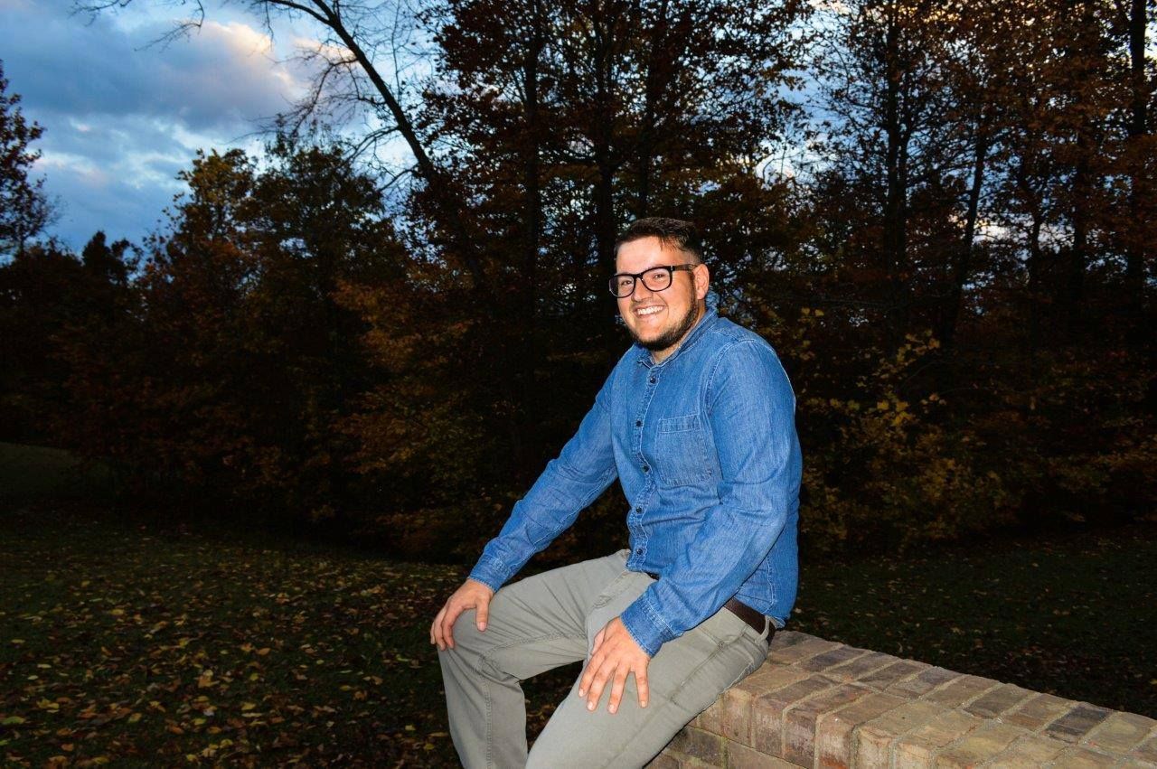 Man in a blue shirt sits on a stone wall at dusk with autumn trees behind him.