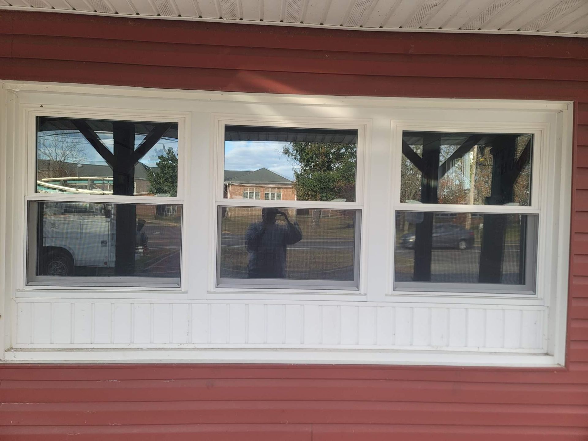 White-trimmed three-pane window set in a red wall, reflecting a street and trees outside.