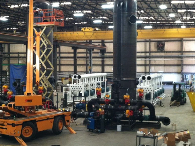 Large industrial equipment inside a factory: tall black structure with connected pipes, orange lift, white machines.