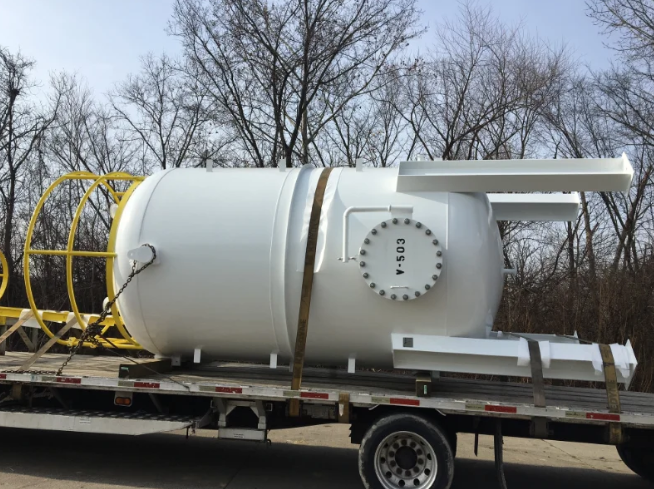 White industrial tank on a flatbed trailer, with a yellow safety cage and arms. Trees in the background.
