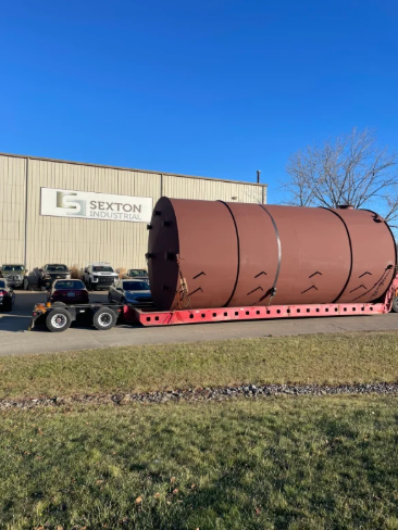 A large brown cylindrical tank on a flatbed trailer in front of Sexton Industries building.