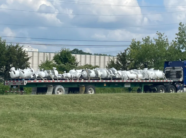 Flatbed truck carrying numerous white cylindrical tanks along a grassy roadside.