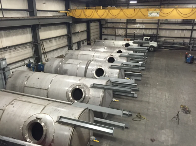 Row of large stainless steel cylindrical tanks in a warehouse, with attached metal supports.