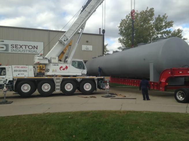 Crane lifting a large, gray industrial tank off a flatbed trailer in front of a building with Sexton Industrial sign.