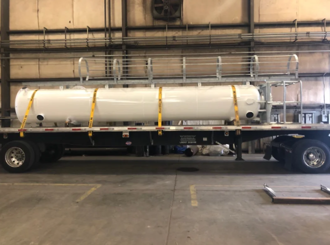 White cylindrical tank secured on a flatbed trailer with yellow straps, in an industrial setting.