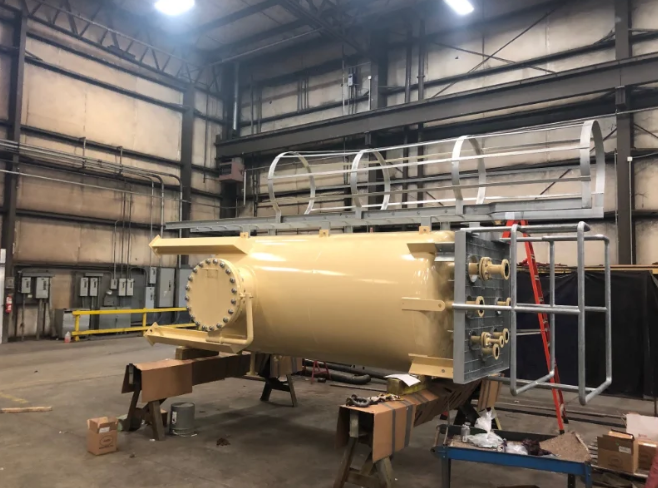 Large, tan industrial tank with metal supports, railing, and ladder, inside a large industrial building.