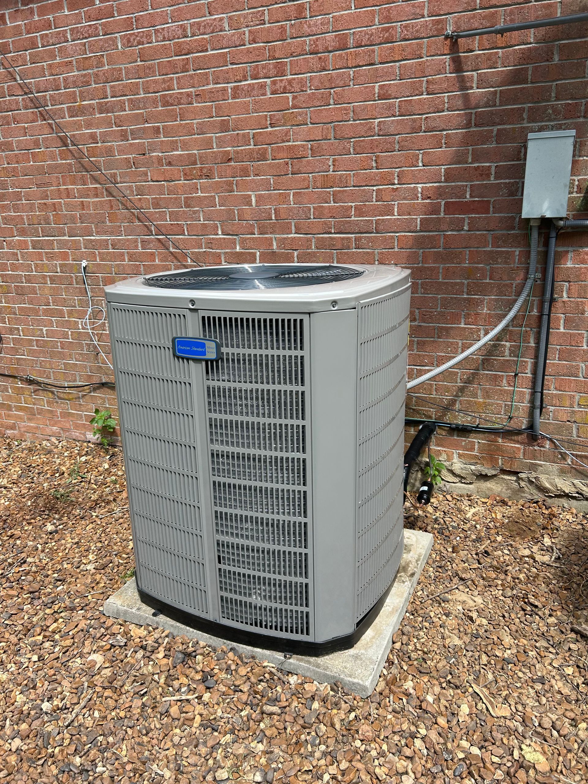 Air conditioner unit against a brick wall. Gray, boxy structure on concrete, surrounded by leaves.