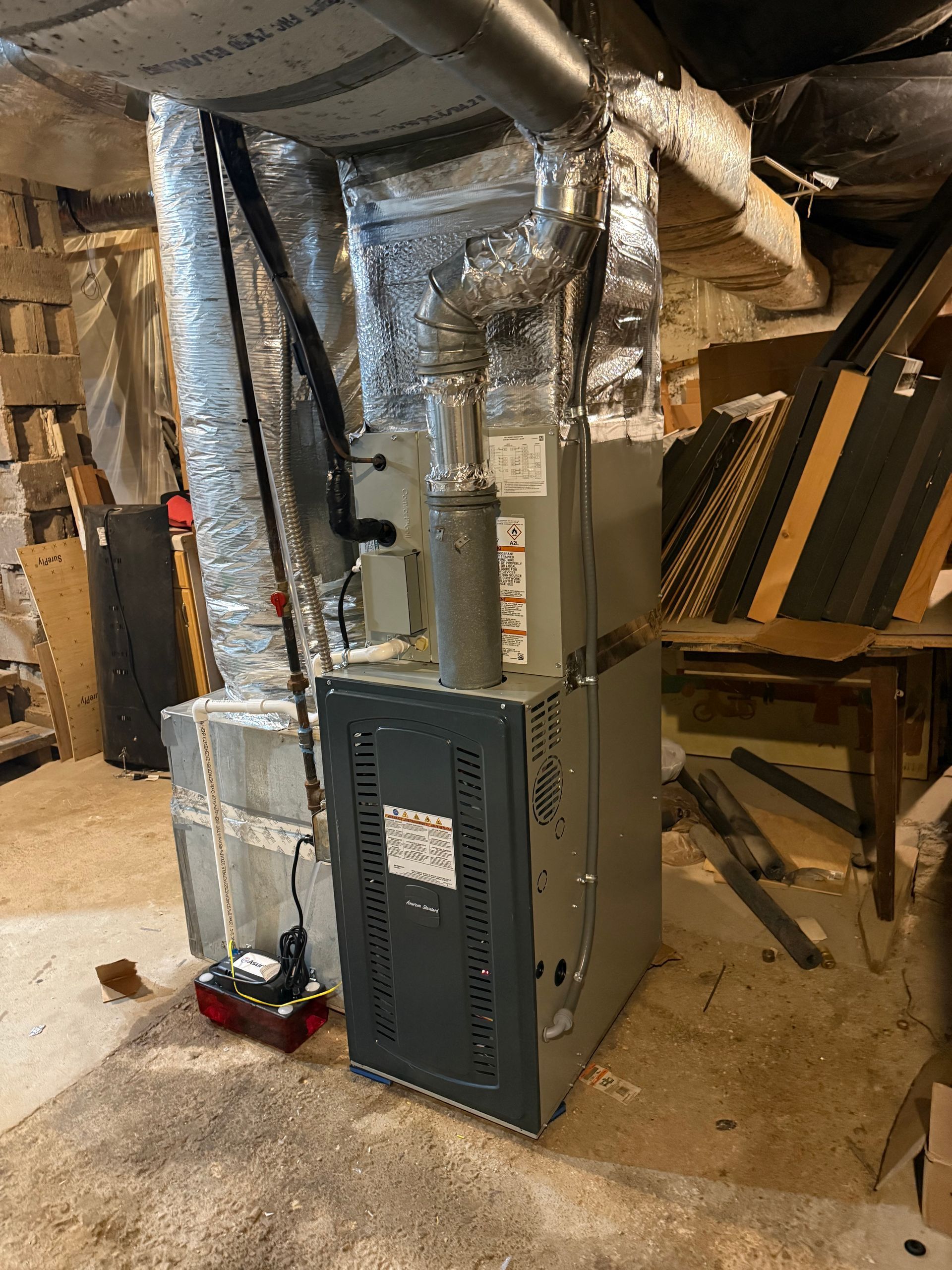 Furnace in basement with ductwork. Gray and silver equipment against stone wall.