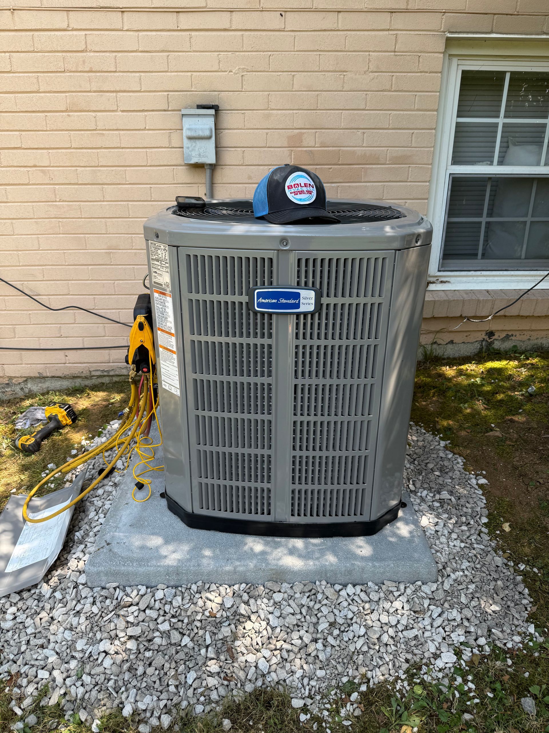 An American Standard air conditioner unit on a gravel base next to a brick wall. A hat sits on top.