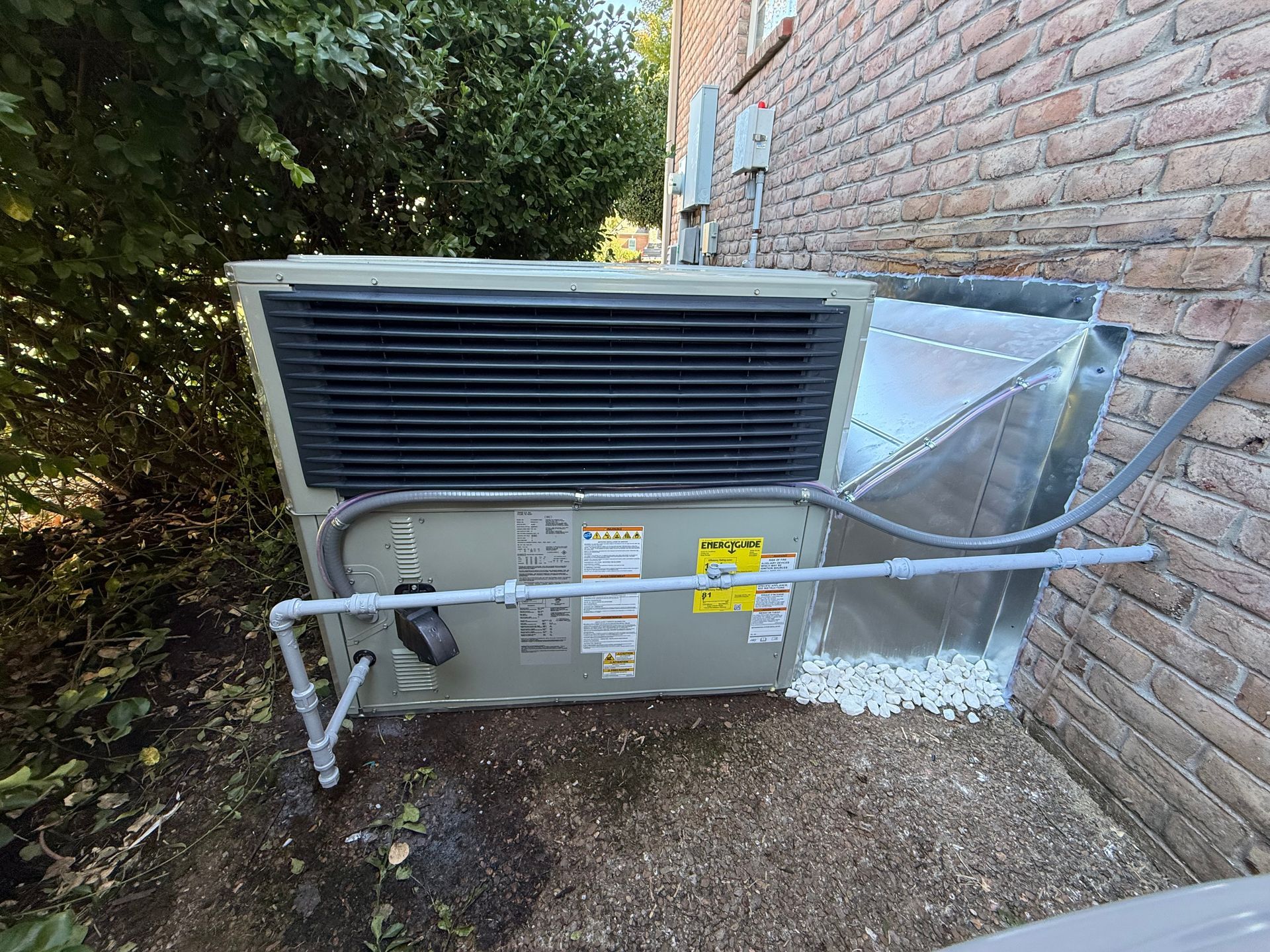 HVAC unit mounted against a brick wall. Gray metal, black grilles, and piping. White gravel base.