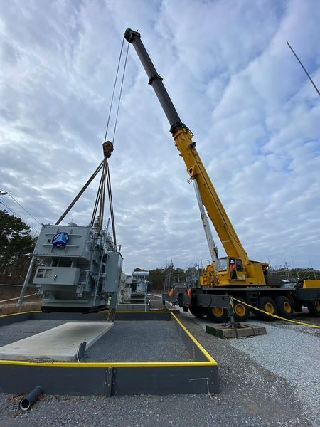 A crane lifts a large electrical transformer at an outdoor power substation, overcast sky above.