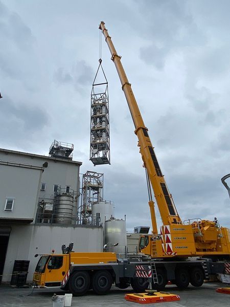 Yellow crane lifting a tall, caged structure at an industrial site on a cloudy day.