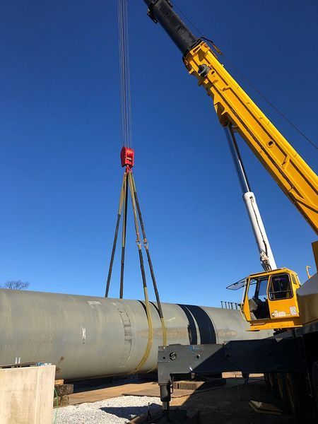 Crane lifting a large cylindrical tank against a blue sky. Yellow crane, red hook, tank is gray.