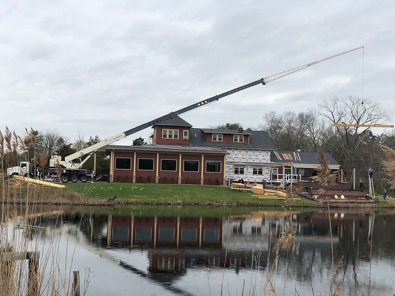 A crane lifts wood beams at a building under construction next to a lake. Overcast sky.