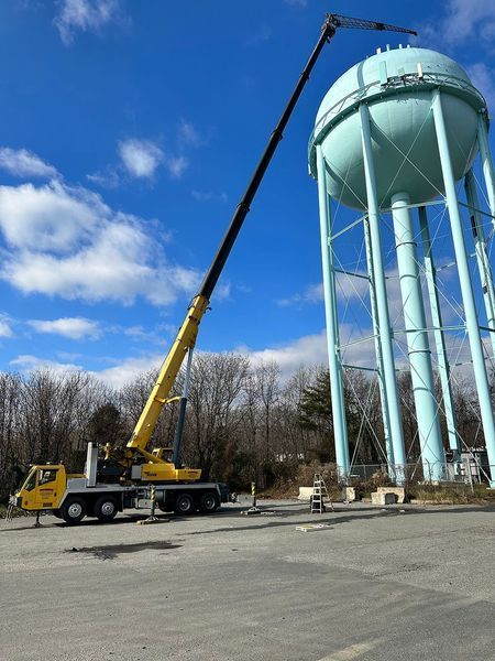 A yellow crane extends towards a tall, light blue water tower against a cloudy sky.