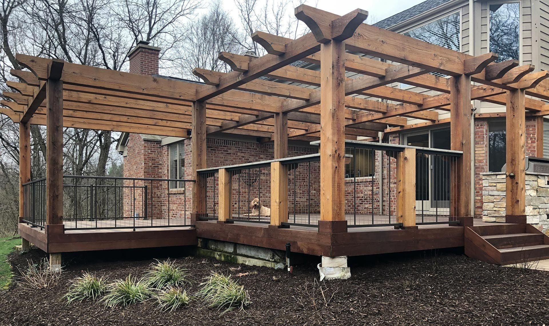 Wooden pergola over a raised deck attached to a brick and stone house.