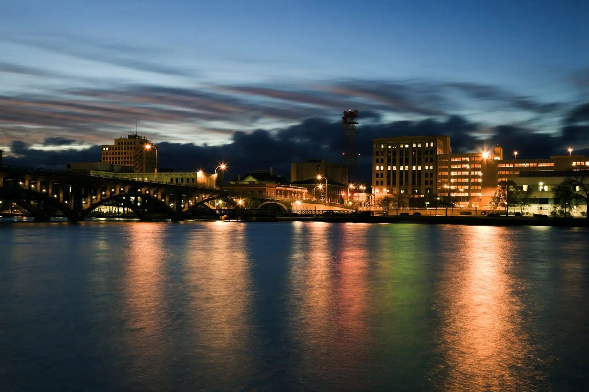 City skyline at dusk reflecting in the water, with a bridge in the foreground. Buildings are illuminated against the dark sky.