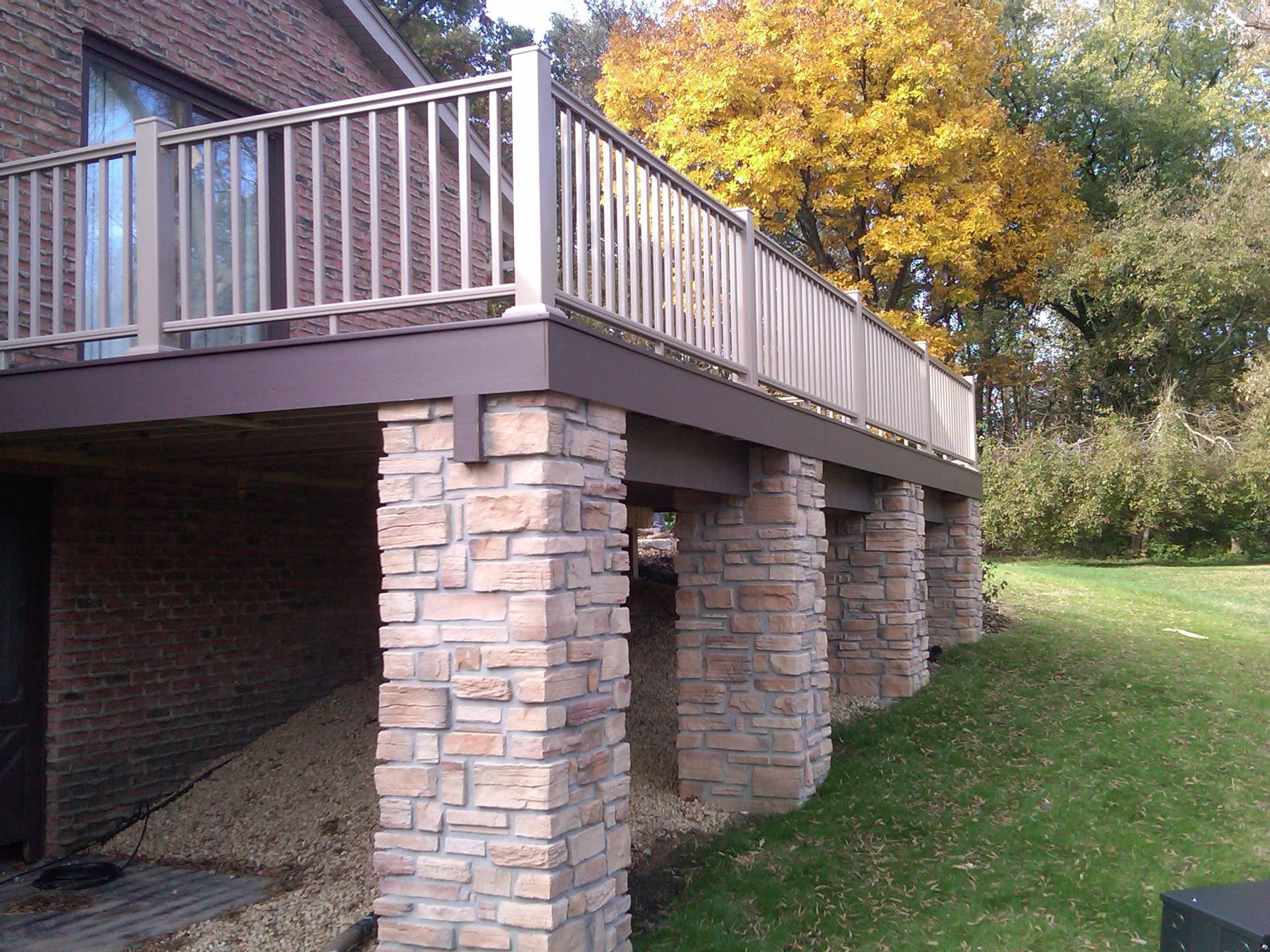 A deck with stone columns on a grassy yard in front of a brick building; fall foliage in background.