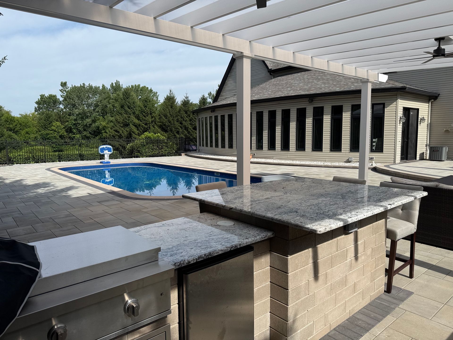 Outdoor kitchen with a grill, bar, and pool with trees in the background.