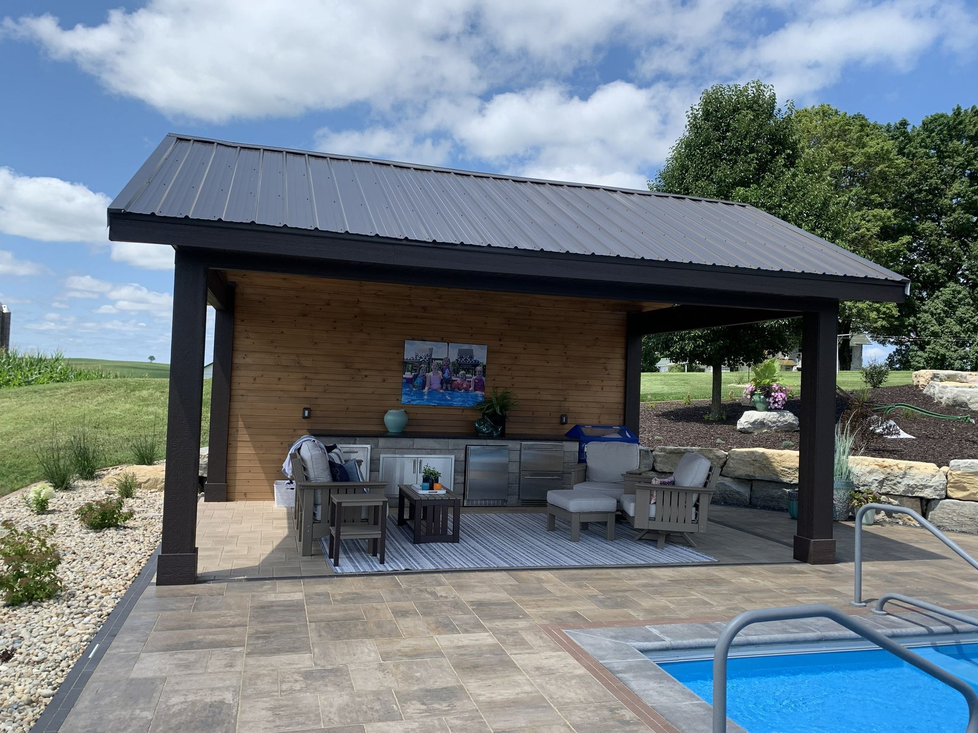 Outdoor kitchen and seating area with a dark metal roof, next to a pool.
