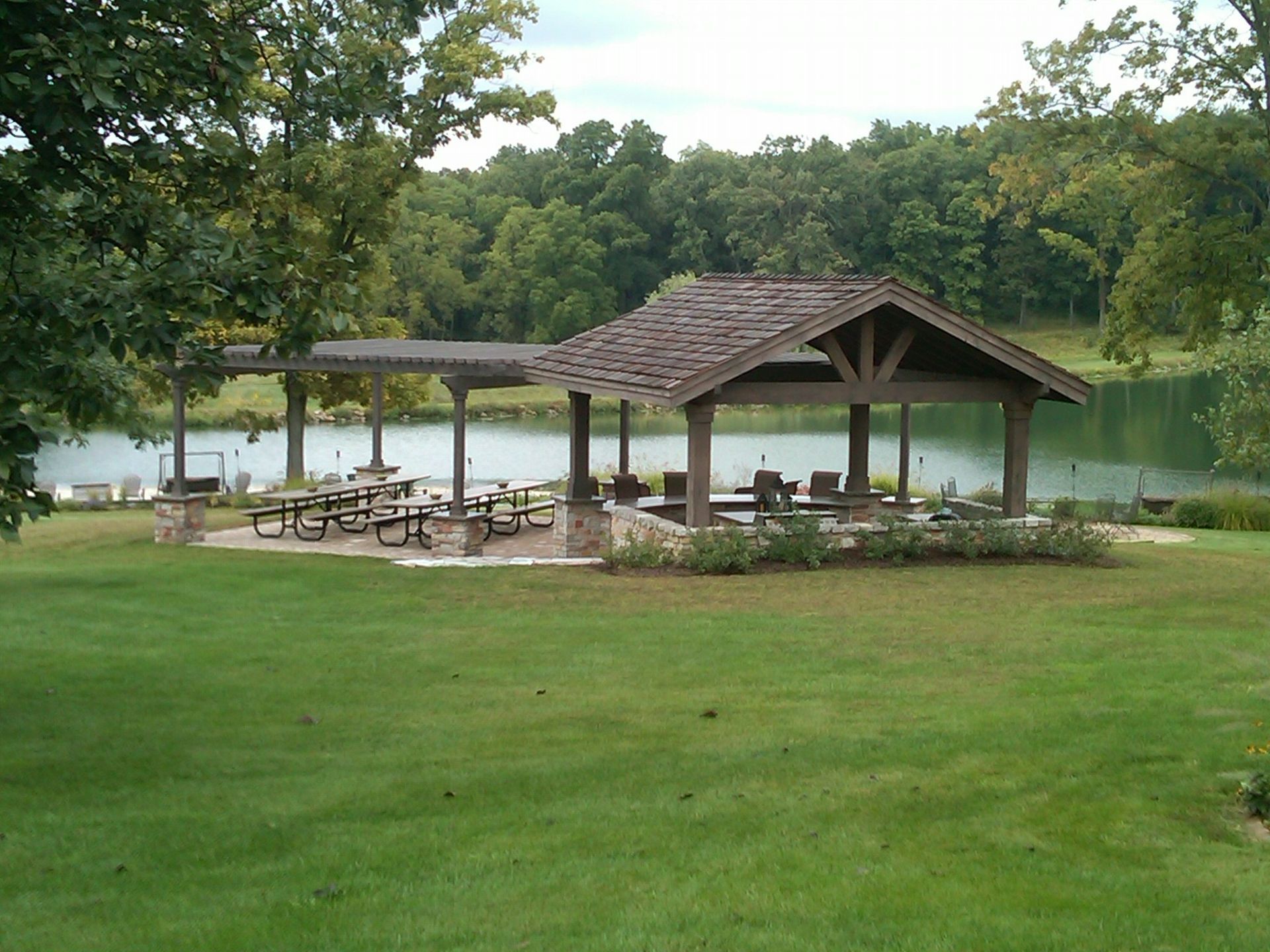 Pavilion with picnic tables overlooking a lake and trees. Green grass in foreground, cloudy sky.