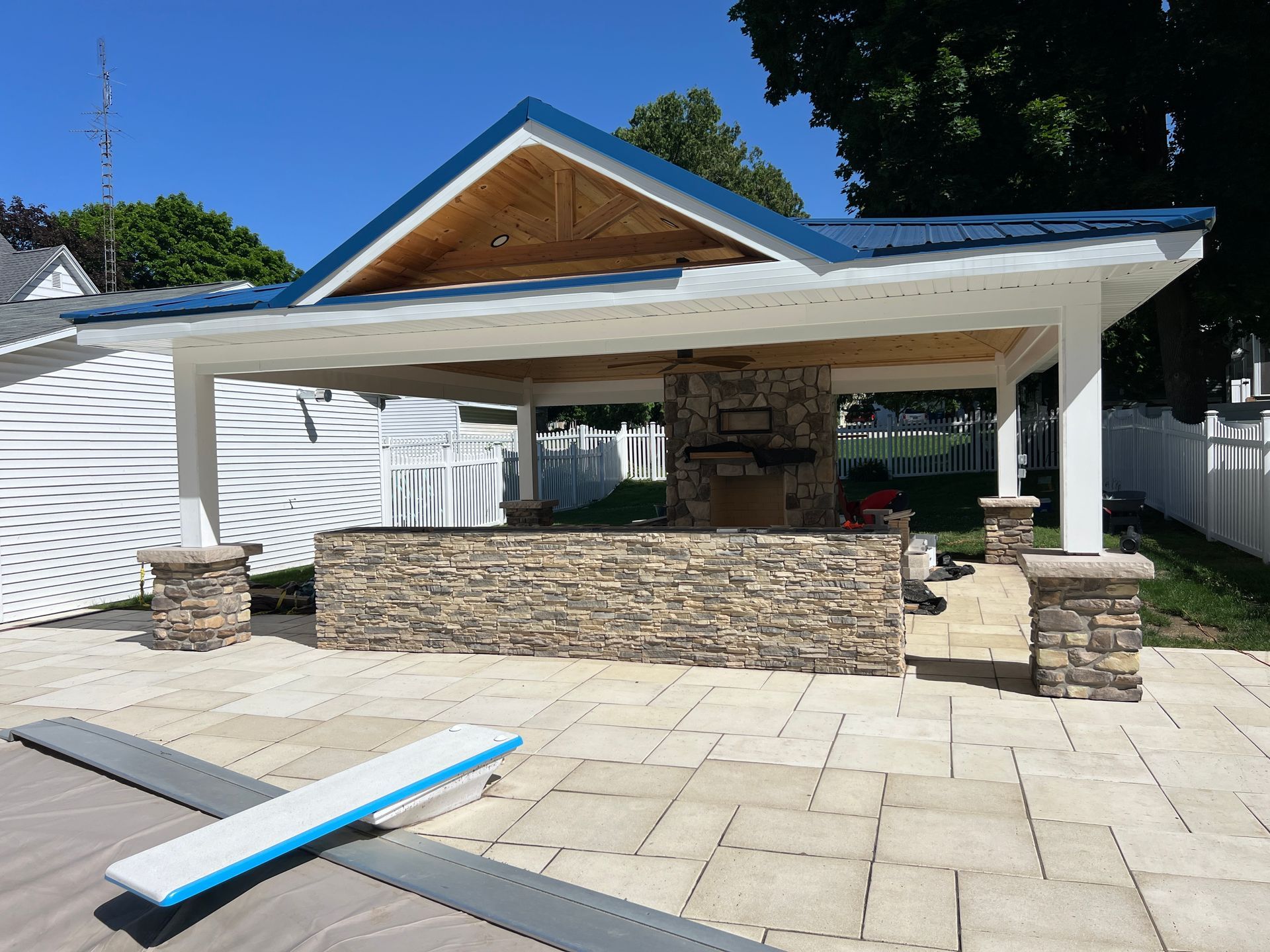 Patio with stone grill and blue-roofed shelter near a swimming pool.